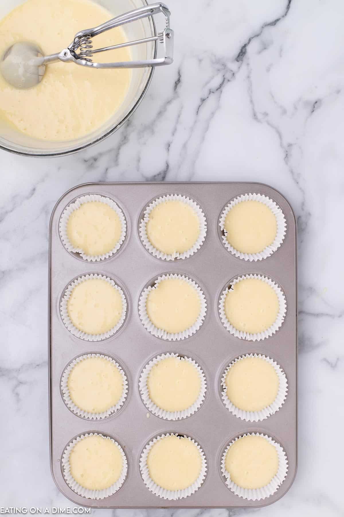 Cupcake batter divided into a cupcake pan lined with cupcake holders with a bowl on the side with cupcake batter and a cookie scoop