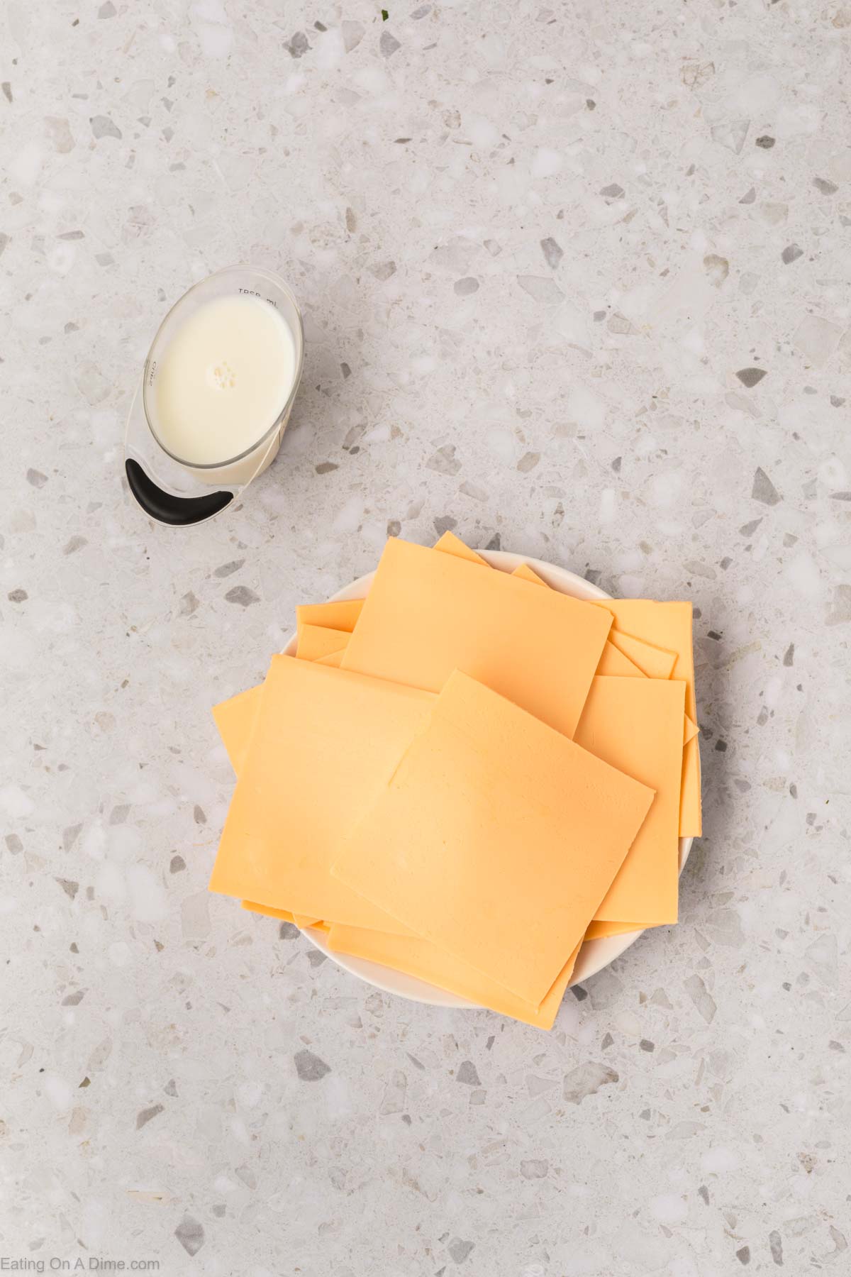 Slices of cheese stacked on a white plate beside a small cup of milk, ready to transform into a creamy American cheese sauce, resting on a speckled gray countertop.