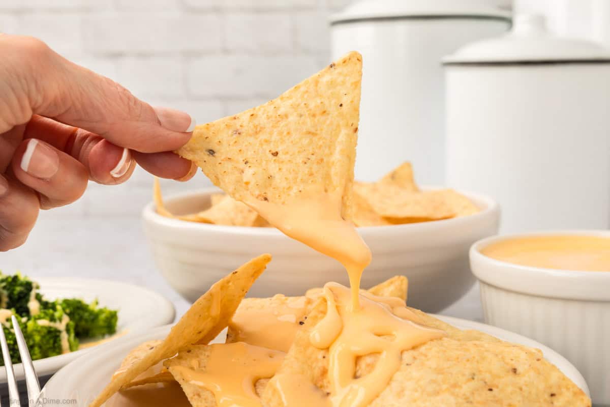 A hand holds a tortilla chip dunked in rich American cheese sauce, its creamy yellow hue stealing the show. Below, a white bowl brims with chips drizzled in more of this cheesy delight. In the background, a smaller bowl of sauce joins a plate adorned with vibrant steamed broccoli.