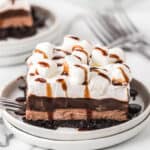 A slice of chocolate layered dessert inspired by a hot chocolate lasagna recipe, topped with whipped cream, mini marshmallows, and a chocolate drizzle on a plate, with a fork beside it and another plate in the background.