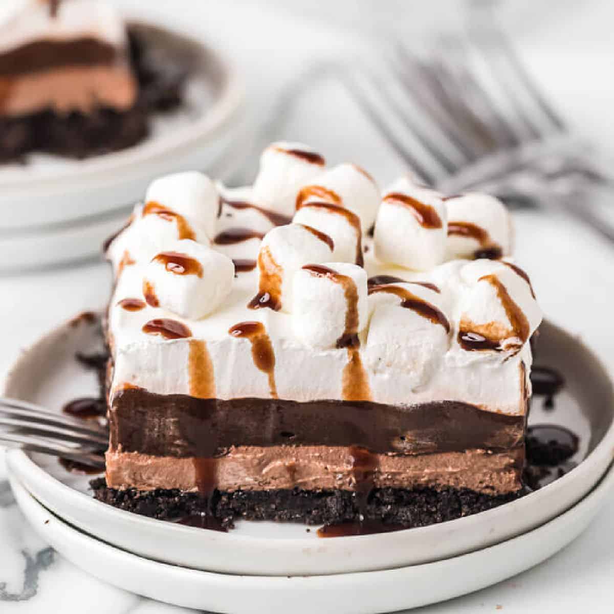 A slice of chocolate layered dessert inspired by a hot chocolate lasagna recipe, topped with whipped cream, mini marshmallows, and a chocolate drizzle on a plate, with a fork beside it and another plate in the background.