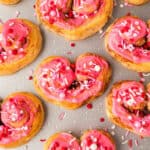 Heart shaped cinnamon rolls topped with pink icing and colorful red, white, and pink sprinkles are arranged on a light surface, with a scattering of icing and sprinkles around the sweet treats.