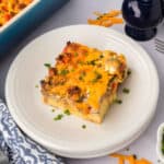 A square slice of Sausage and Egg Breakfast Casserole topped with melted cheddar cheese and chopped chives sits on a white plate. A casserole dish, fork, and napkin are nearby.