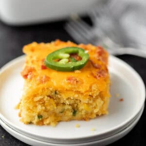 A slice of cheesy Jalapeño Popper corn Casserole topped with sliced jalapeño, served on a white plate with a fork and napkin in the background.