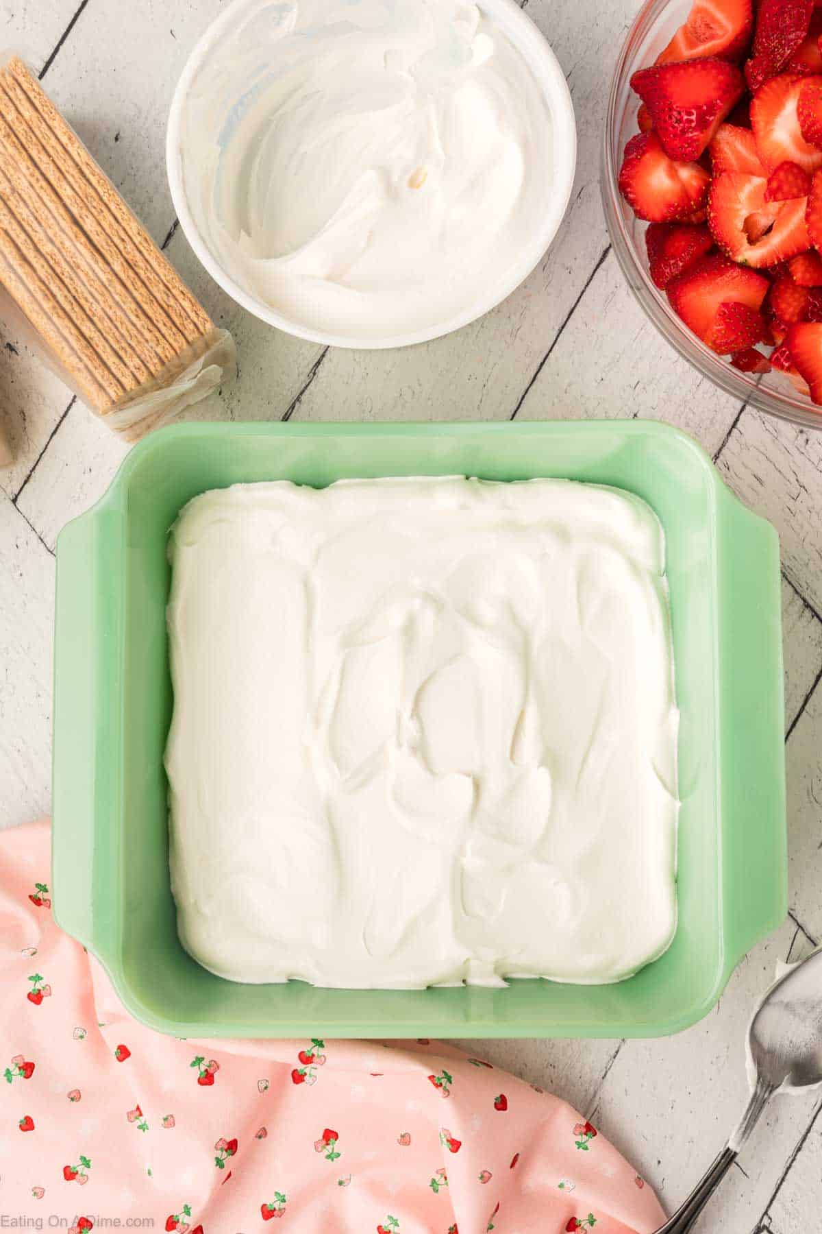 Spreading cool whip in the bottom of the baking dish with a bowl of fresh strawberries on the side with cool whip and graham crackers