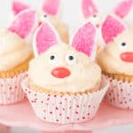 Easter Bunny Cupcakes decorated with white frosting, candy eyes, pink sugar ears, and red candy noses to look like bunnies, displayed on a pink cake stand.