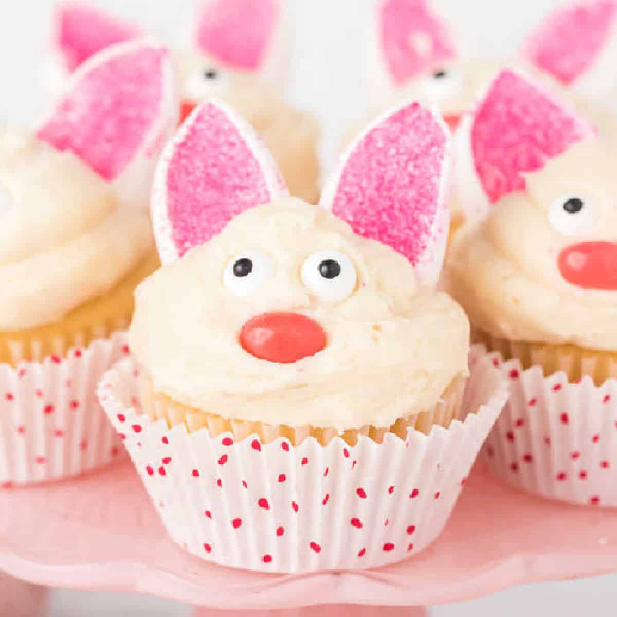 Easter Bunny Cupcakes decorated with white frosting, candy eyes, pink sugar ears, and red candy noses to look like bunnies, displayed on a pink cake stand.