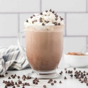 A glass mug filled with iced mocha, inspired by the Starbucks hot chocolate recipe, topped with whipped cream and chocolate chips, surrounded by scattered chips on a white surface with a striped towel and a small bowl in the background.