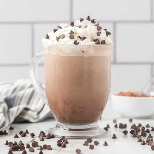 A glass mug filled with iced mocha, inspired by the Starbucks hot chocolate recipe, topped with whipped cream and chocolate chips, surrounded by scattered chips on a white surface with a striped towel and a small bowl in the background.