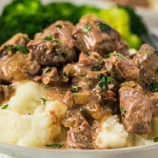 A plate of creamy mashed potatoes topped with Instant Pot Beef Tips and Gravy, garnished with chopped herbs, with steamed broccoli in the background.