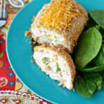 A breaded, baked chicken breast stuffed with creamy filling and chopped green peppers sits on a blue plate next to fresh spinach leaves—an irresistible Jalapeño Popper Stuffed Chicken Breasts Recipe on a colorful patterned tablecloth.