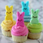 Four Peeps Cupcakes with colorful frosting—yellow, pink, green, and blue—each topped with a matching marshmallow bunny Peep, displayed on a white marble surface.