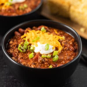 A black bowl of chili from an Easy Chili Recipe, topped with shredded cheddar cheese, sour cream, and chopped green onions, with another bowl and cornbread visible in the background.