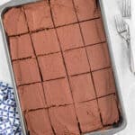 A rectangular baking pan filled with an easy chocolate cake recipe, topped with rich frosting and cut into 20 even squares, sits beside two forks and a blue-and-white patterned napkin on a light surface.