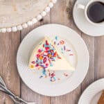 A slice of white frosted cake made from the Best Vanilla Cake Recipe, topped with colorful sprinkles on a white plate, sits next to a cup of black coffee on a rustic wooden table.
