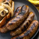 Grilled beer brats with browned grill marks on a dark plate, with a sandwich bun and sautéed onions partially visible in the corner.