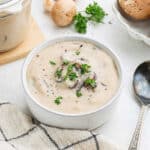 A bowl of Homemade Cream of Mushroom Soup Recipe sits garnished with chopped parsley, sliced mushrooms visible. A spoon rests nearby, with fresh mushrooms and parsley scattered on the table in the background.