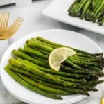 A white plate with roasted air fryer asparagus spears garnished with a slice of lemon. In the background, a white platter holds more asparagus, and two gold forks are visible on the side.