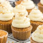 Several carrot cupcakes with cream cheese frosting, made with an easy carrot cake cupcakes recipe, are topped with chopped walnuts and arranged on a cooling rack. The cupcakes are in brown paper liners and look moist and freshly baked.