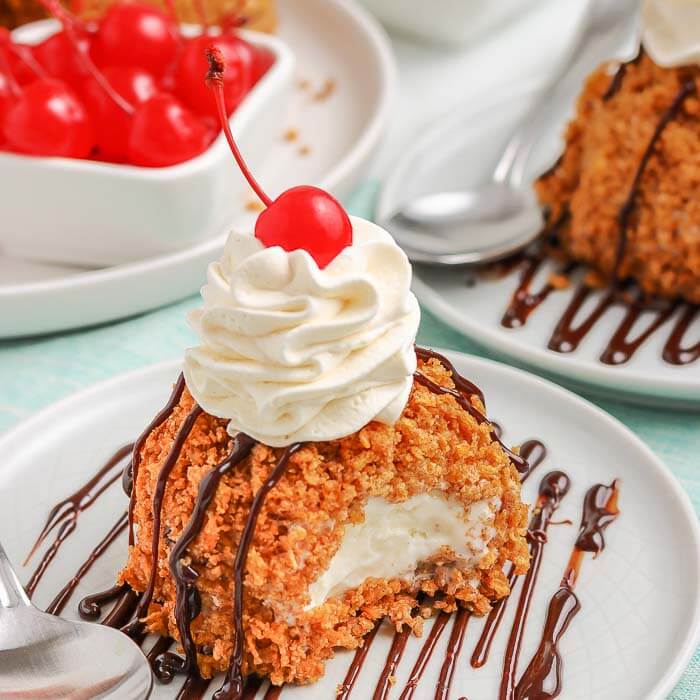 Fried Ice Cream on a white plate topped with whipped cream and a cherry with more cherries behind it 