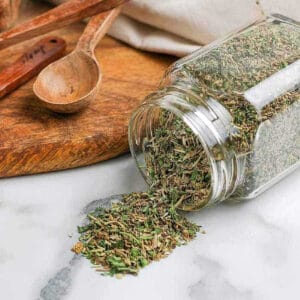 A glass jar tipped over on a marble countertop, spilling dried mixed herbs. A wooden spoon rests nearby on a round wooden cutting board, with a cream-colored cloth in the background. The scene evokes the preparation of an Italian seasoning recipe in a cozy kitchen setting.