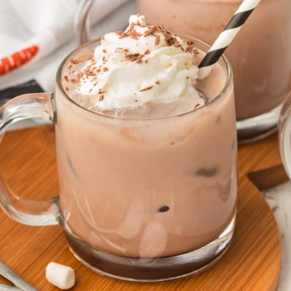 A glass mug filled with creamy iced chocolate milk, topped with whipped cream and chocolate shavings, and a black-and-white striped strawโthis is the perfect presentation for your favorite iced hot chocolate recipe. Another similar drink sits beside it on a wooden surface.
