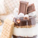 A jar filled with layers of chocolate, crushed graham crackers, and marshmallow, topped with mini marshmallows and two pieces of Hershey's chocolate—this S'mores Parfait Recipe is finished with a graham cracker leaning against the jar.