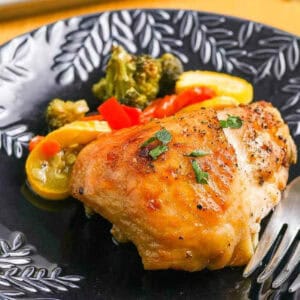 A black plate with a serving of sheet pan chicken thigh dinner garnished with herbs, accompanied by cooked broccoli, yellow squash, and red bell peppers. A fork rests beside the food.