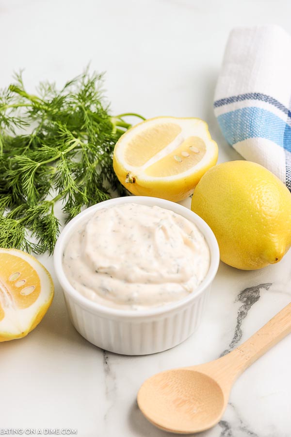Tartar sauce in a white bowl with lemons and dill next to it. 