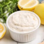 A small white ramekin filled with creamy dill sauce, inspired by the Red Lobster Tartar Sauce Recipe, sits on a marble surface, surrounded by fresh lemons and dill.