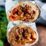 Two beef burritos wrapped in foil, cut in half to reveal a delicious beef burrito recipe with ground beef, rice, tomatoes, onions, and cheese, stacked on top of each other on a wooden surface.