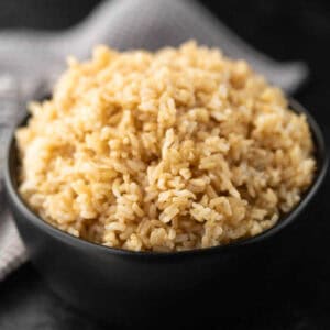 A black bowl filled with fluffy Instant Pot Brown Rice sits on a dark surface, with a blurred checkered cloth in the background.
