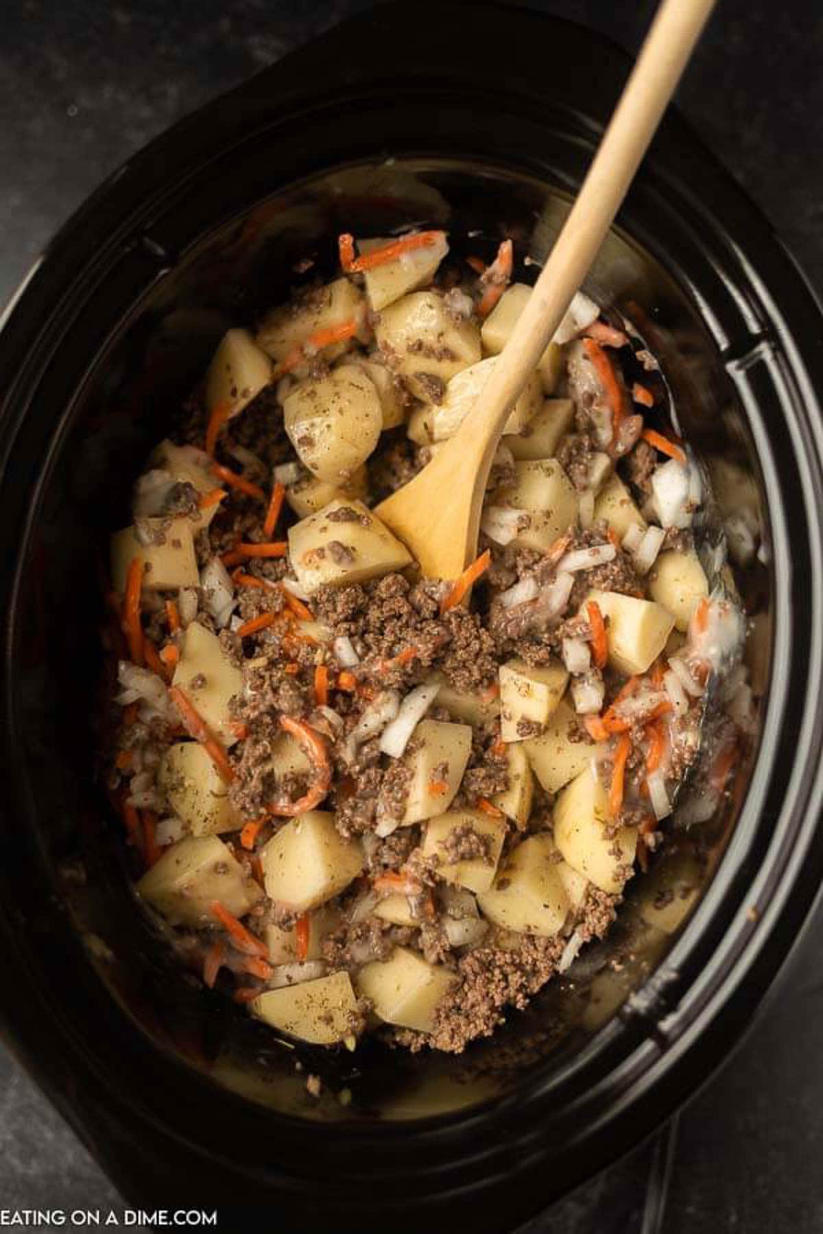 The browned ground beef, potatoes, onions, carrots, seasoning, beef broth and cream of mushroom soup stirred together in a crock pot with a wooden spoon.  