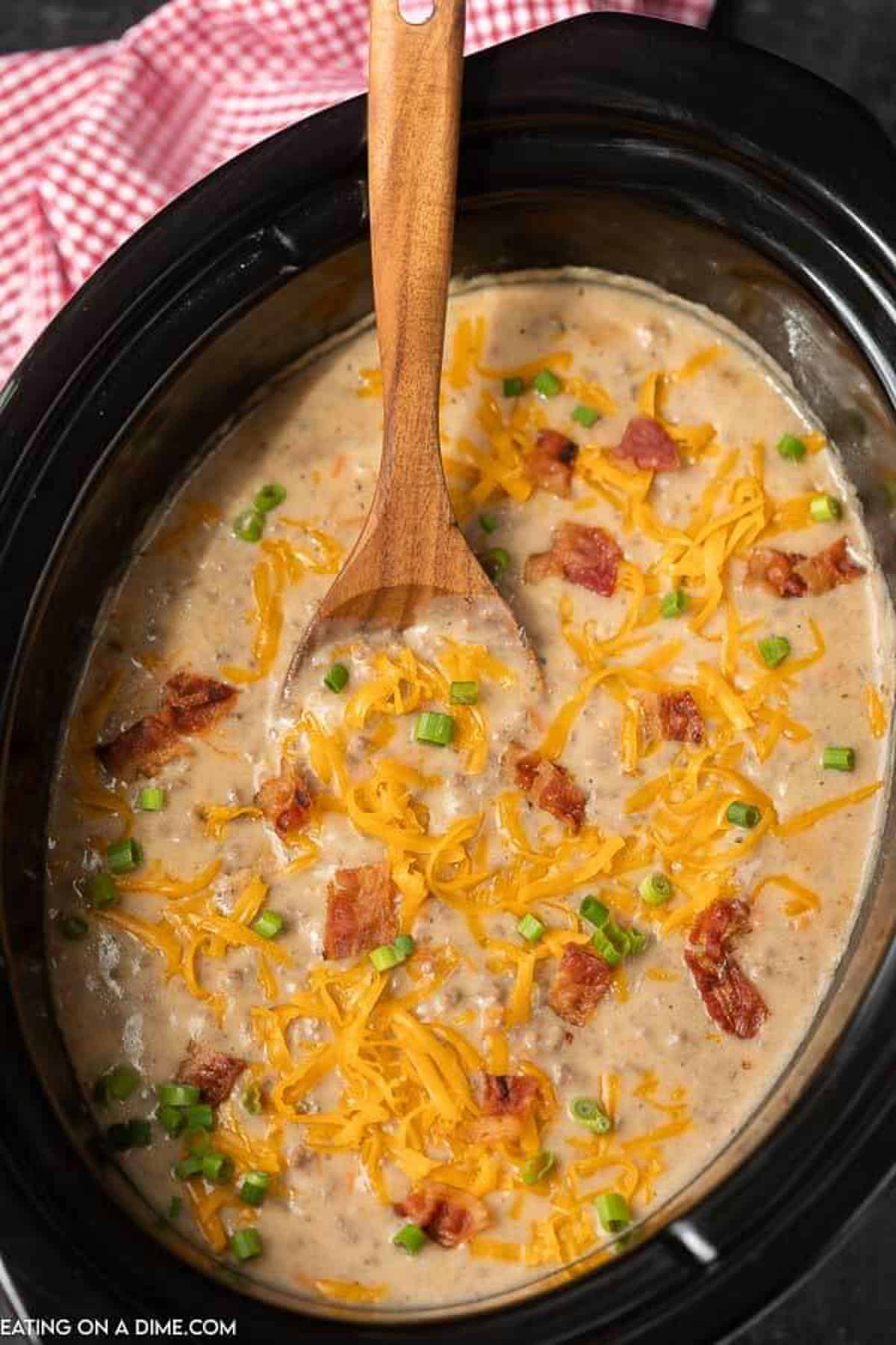 Crock Pot Cheeseburger Soup in a Crock Pot topped with shredded cheese, bacon and diced green onions with a wooden spoon in the crock pot.  
