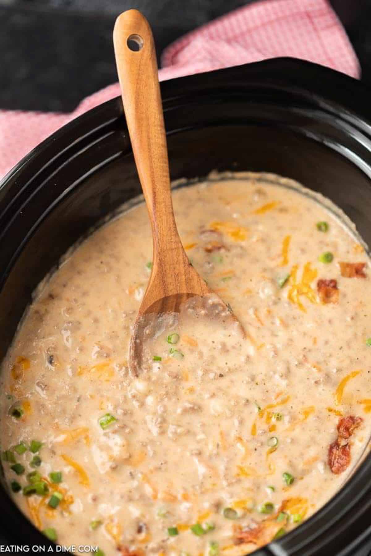 Crock Pot Cheeseburger Soup in a Crock Pot topped with shredded cheese, bacon and diced green onions with a wooden spoon in the crock pot.  