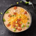 A bowl of creamy Crockpot Cheeseburger Soup Recipe topped with shredded cheddar cheese, crispy bacon pieces, and chopped green onions sits on a dark surface, with a spoon in the background.