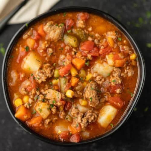 A bowl of hearty Crockpot Vegetable Beef Soup filled with ground beef, potatoes, carrots, corn, green beans, and tomatoes in a rich, red broth, garnished with fresh herbs.