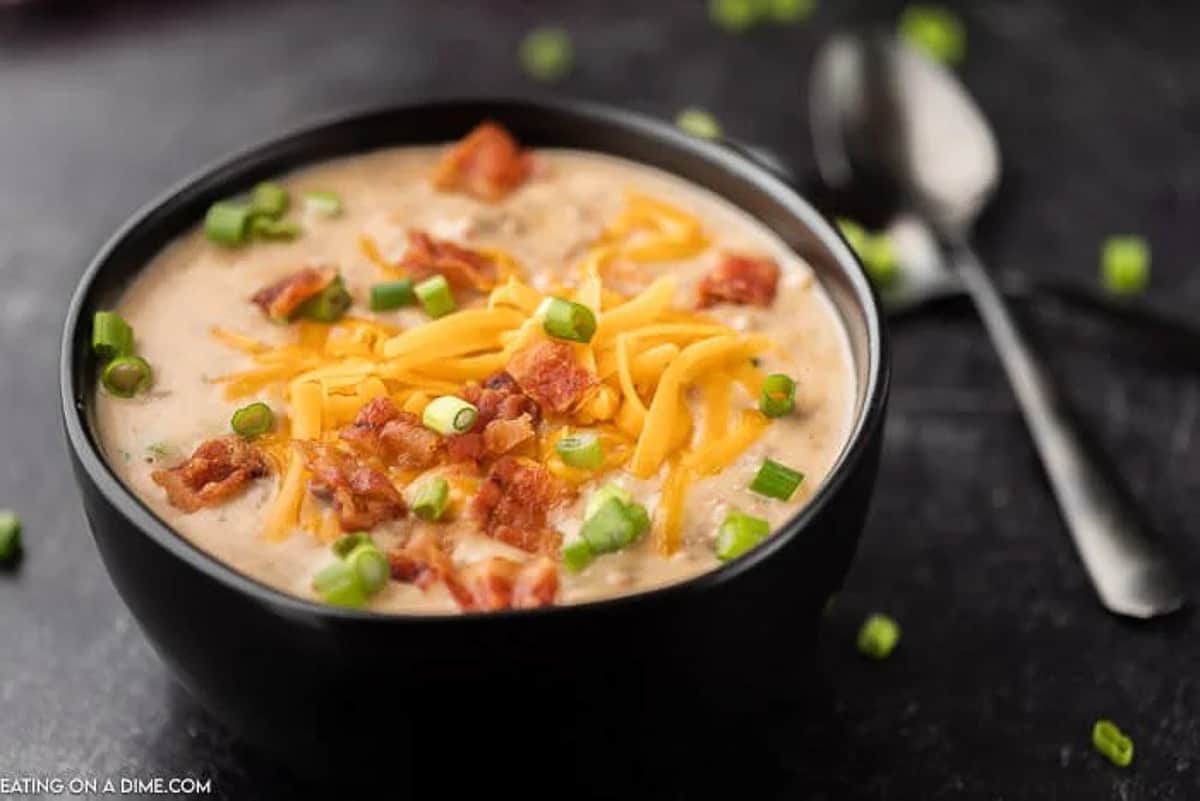 Crock Pot Cheeseburger soup in a black bowl topped with shredded cheddar cheese, diced bacon and diced green onions. 

