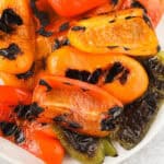 A close-up of grilled bell peppers in red, orange, and green hues shows char marks and slightly wrinkled skin on a white plate, illustrating how to grill bell peppers for vibrant color and flavor.