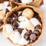 A close-up of a waffle cone filled with melted chocolate chips and toasted mini marshmallows, these s'mores campfire cones perfectly capture the classic dessert in a fun, portable treat.