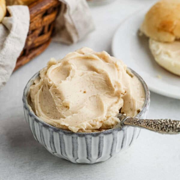 A small, textured ceramic bowl filled with whipped cinnamon butter, inspired by the famous Texas Roadhouse Butter Recipe, with a decorative silver spoon resting inside. In the background, there is a basket of bread and a plate with a roll.