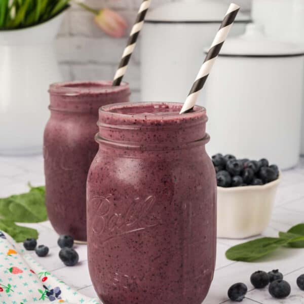Two mason jars filled with vibrant purple blueberry spinach smoothie recipe, each with a black-and-white striped straw. Fresh blueberries, spinach leaves, and a white bowl of blueberries are nearby on a bright, light surface.