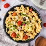 A bowl of Easy Pasta Salad with rotini, cherry tomatoes, cucumber, red onion, black olives, cheese cubes, and herbs sits on a table, surrounded by plates, utensils, and fresh vegetables.
