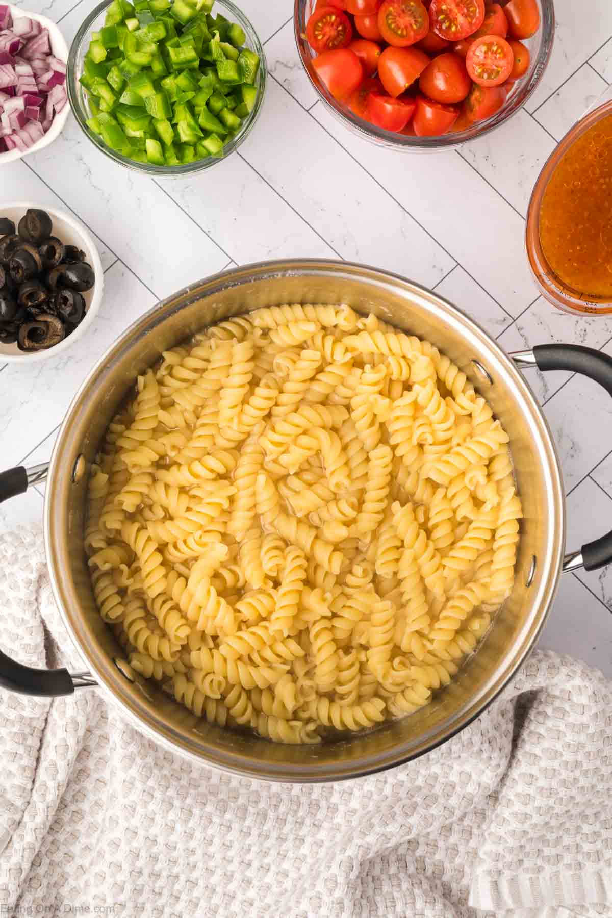 Cooking Rotini Noodles in a large pot of water with bowls of cherry tomatoes, chopped bell peppers, red onions, and black olives on the side