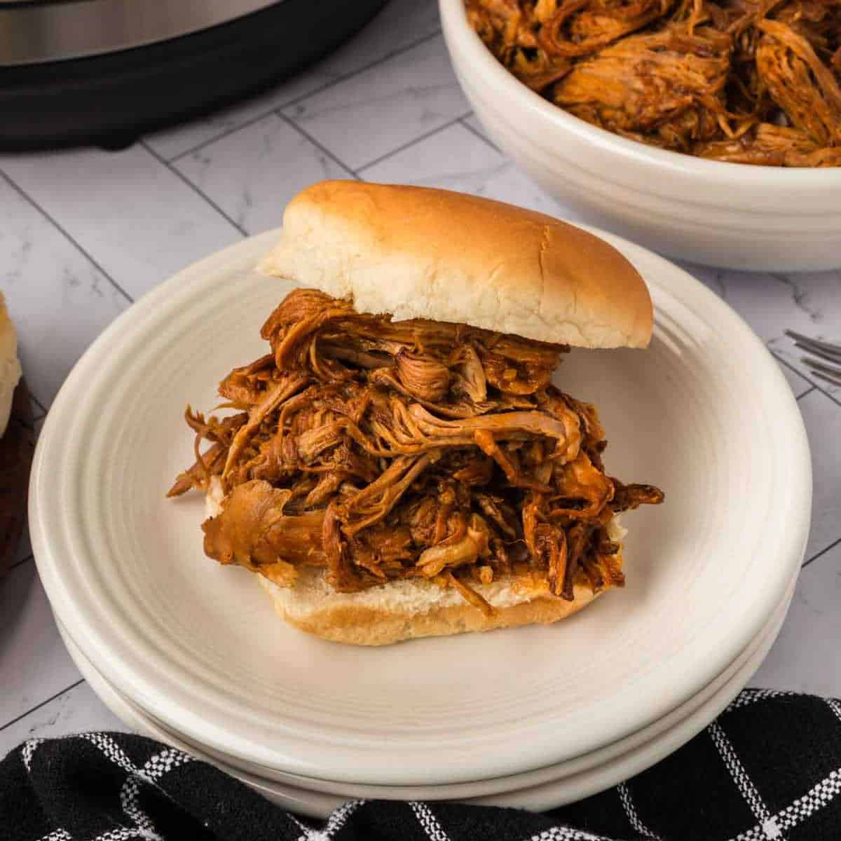 A pulled chicken sandwich on a white bun sits on a white plate, with a bowl of shredded chicken and Instant Pot Pulled Pork in the background, and a black-and-white striped napkin nearby.