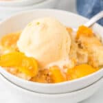 A bowl of peach cobbler made from a crock pot peach cobbler recipe, topped with a scoop of vanilla ice cream, with a spoon beside the dessert.