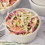 A bowl of coleslaw salad with a fork and a white bowl. extra coleslaw and bowls in the background.