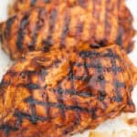 Two grilled chicken breasts with dark grill marks, coated in a glossy barbecue sauce, shown close-up on a white surface