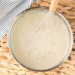 A metal saucepan filled with creamy white sauce is being whisked with a metal whisk—a perfect step if you're learning how to make a roux. The pan rests on a woven placemat next to a striped cloth.