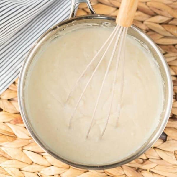 A metal saucepan filled with creamy white sauce is being whisked with a metal whisk—a perfect step if you're learning how to make a roux. The pan rests on a woven placemat next to a striped cloth.